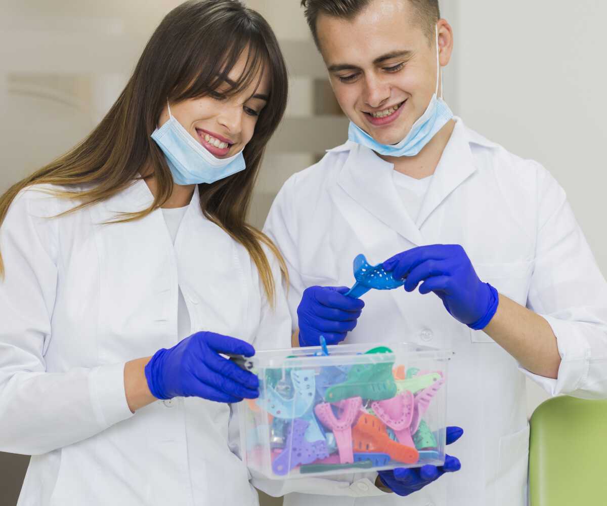 Two dentists smiling while holding a box of colorful dental retainers, both wearing white lab coats, blue gloves, and face masks around their necks in a dental clinic.