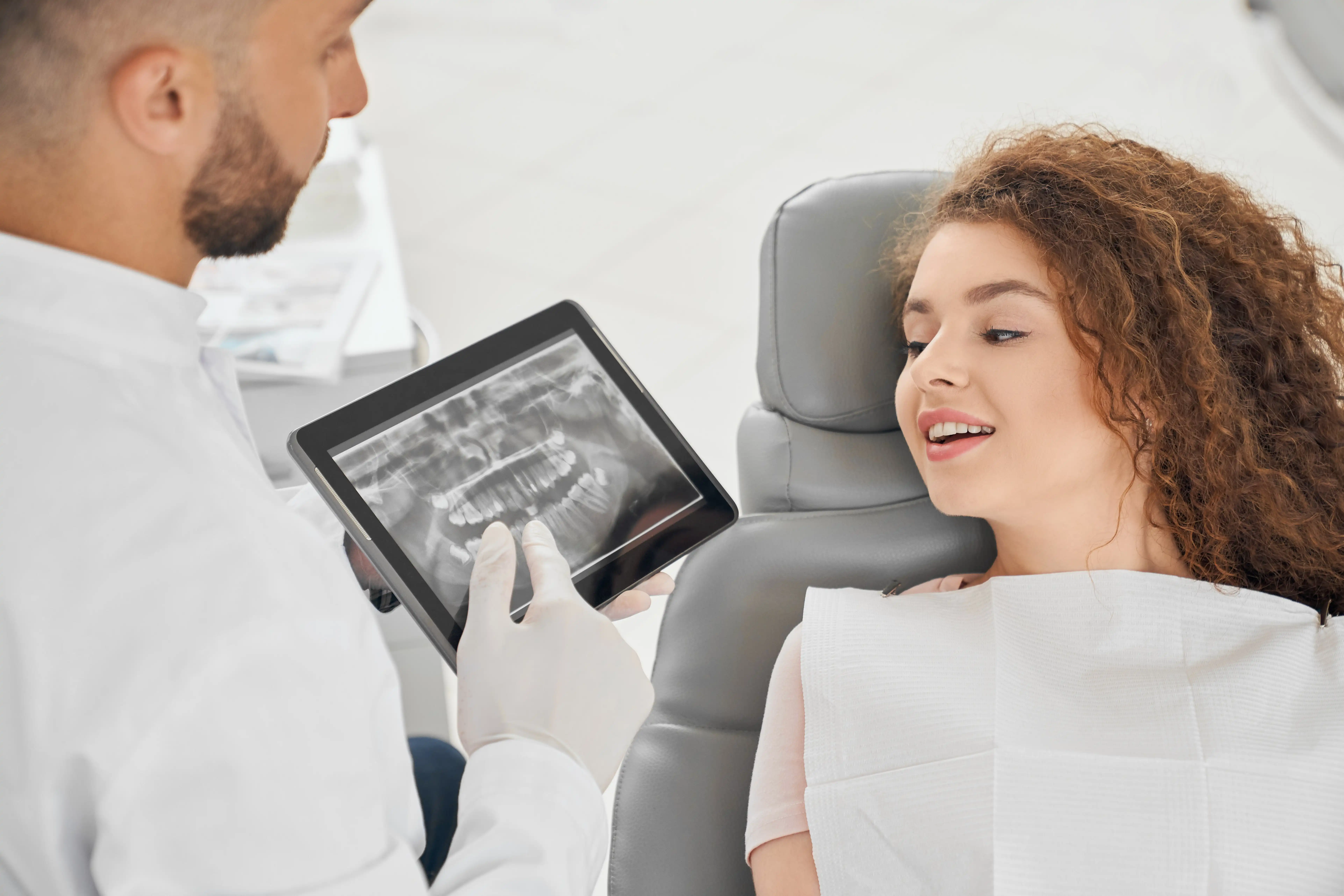 Bir diş hekimi, kadın bir hastaya dijital dental x-ray görüntüsünü tablet üzerinden gösteriyor.